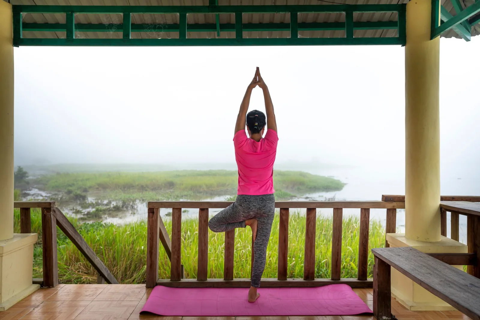 Back view full body of anonymous fit female with raised arms practicing Tree pose while doing yoga on veranda in tropical country