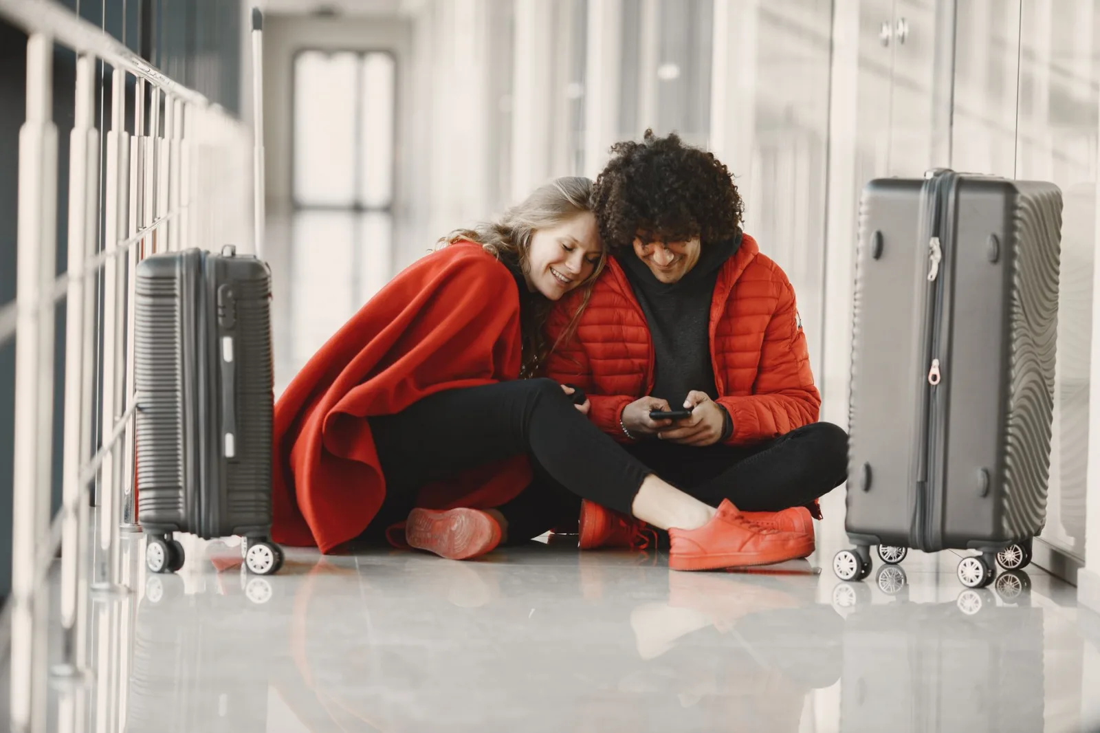 Happy couple sitting on the airport floor with luggage, sharing a moment using smartphone.