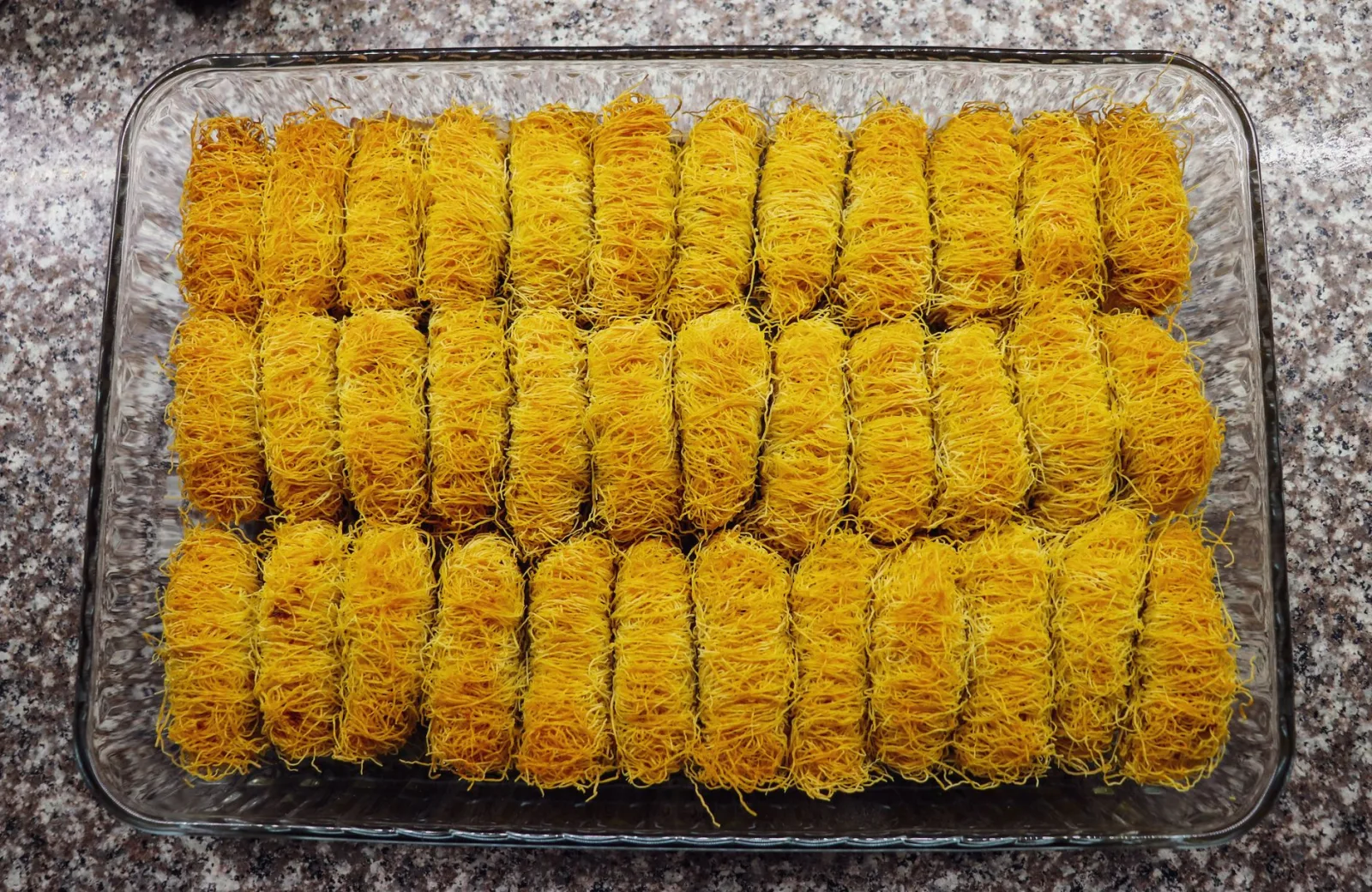 Glass tray filled with golden kataifi pastry rolls on a granite countertop, ready for baking.