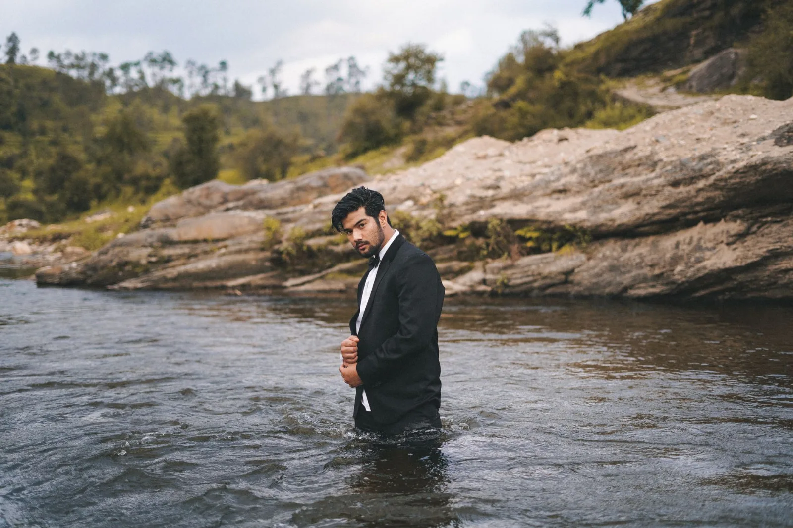 A fashionable man in a suit poses confidently in a river with a serene natural backdrop.