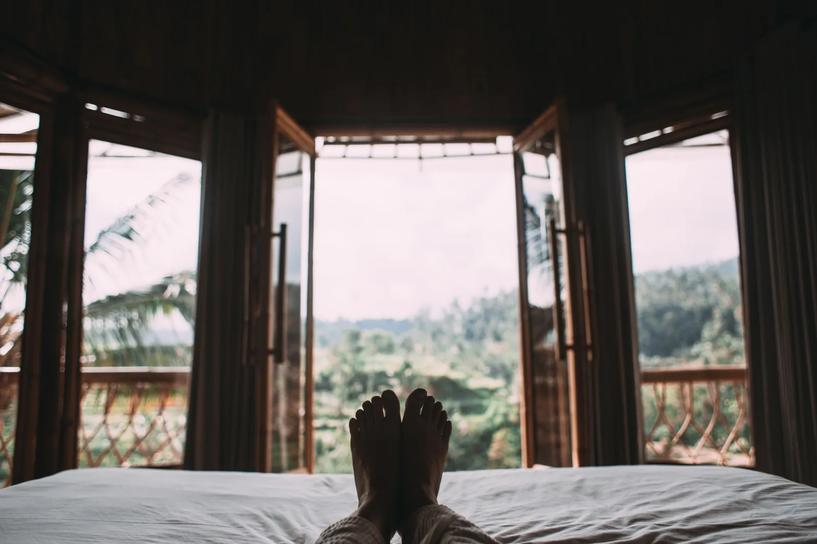 Crop person chilling on white bed in dark cozy room with balcony and wide windows in exotic country in daytime