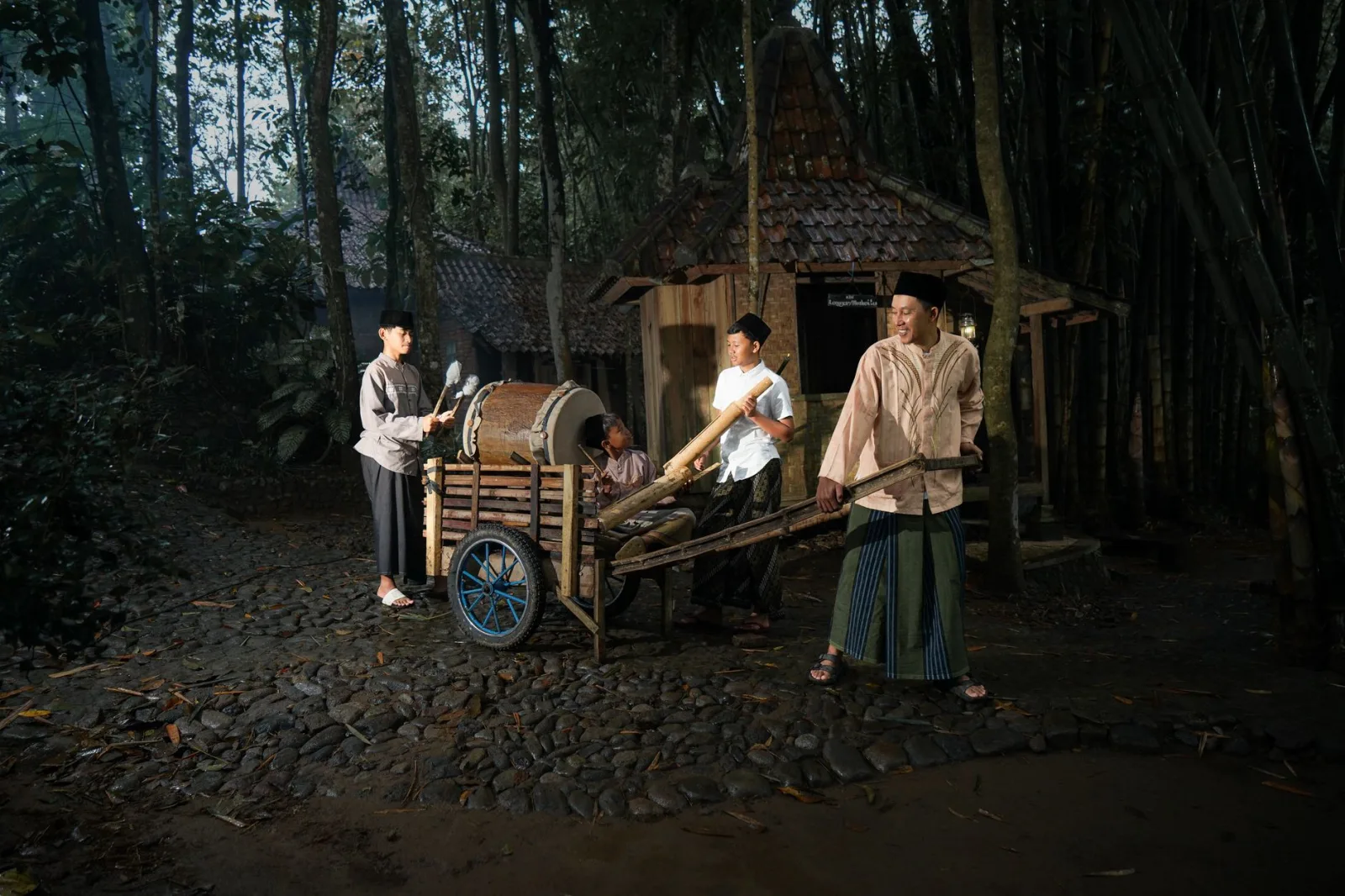 Four men in traditional Indonesian attire playing a drum outdoors with a joyful expression.