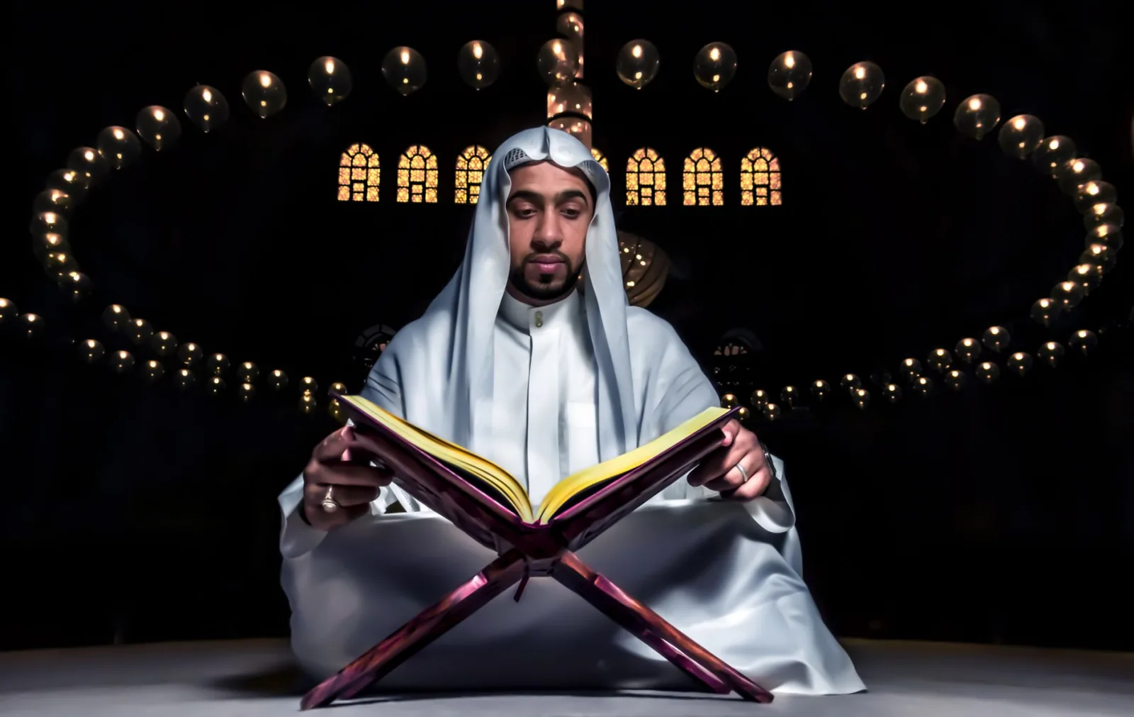 Middle Eastern man in traditional attire reading a holy book inside a mosque with ambient lighting.