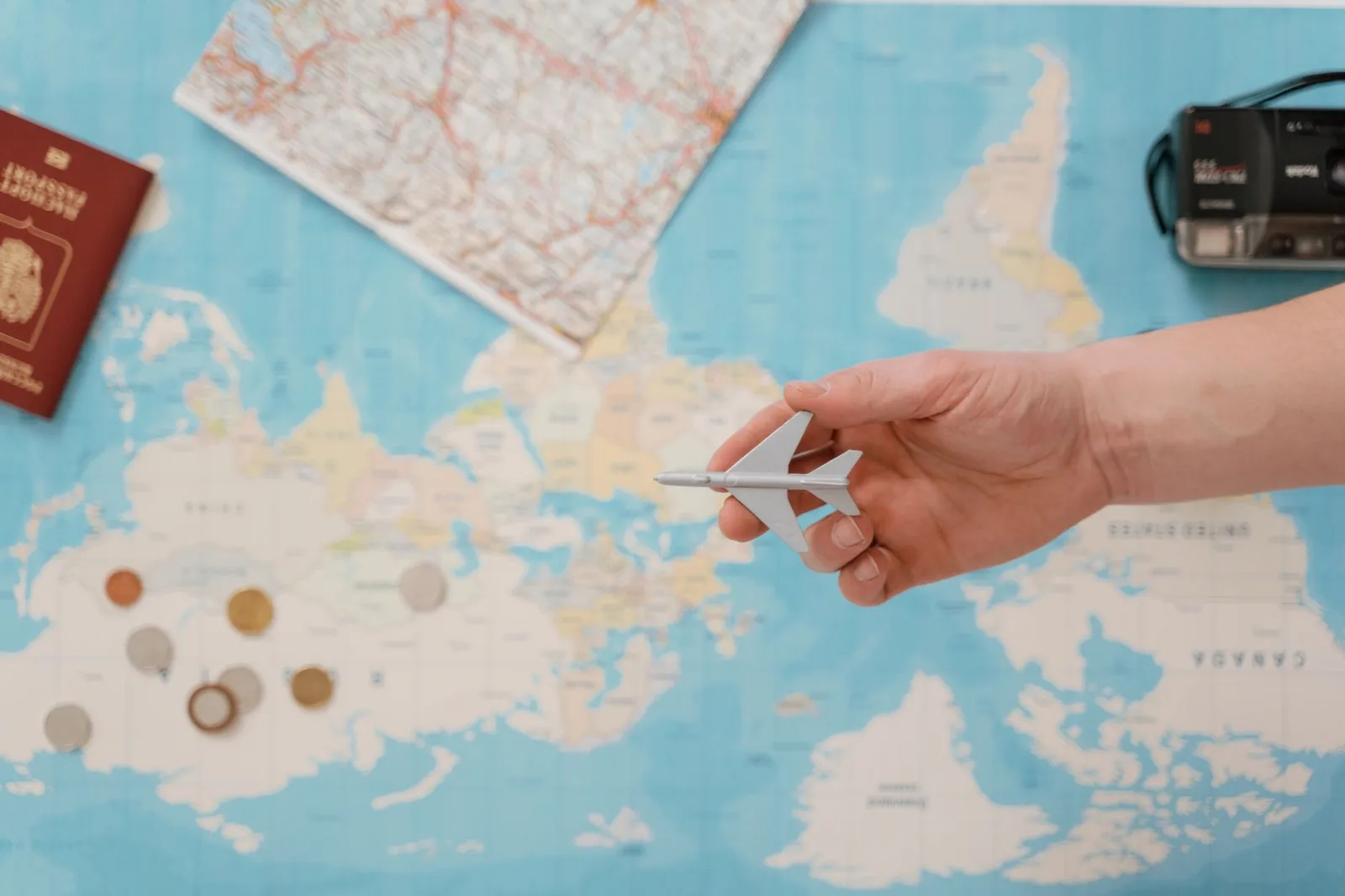 Hand holding a toy airplane over a world map, surrounded by coins, camera, and passport.