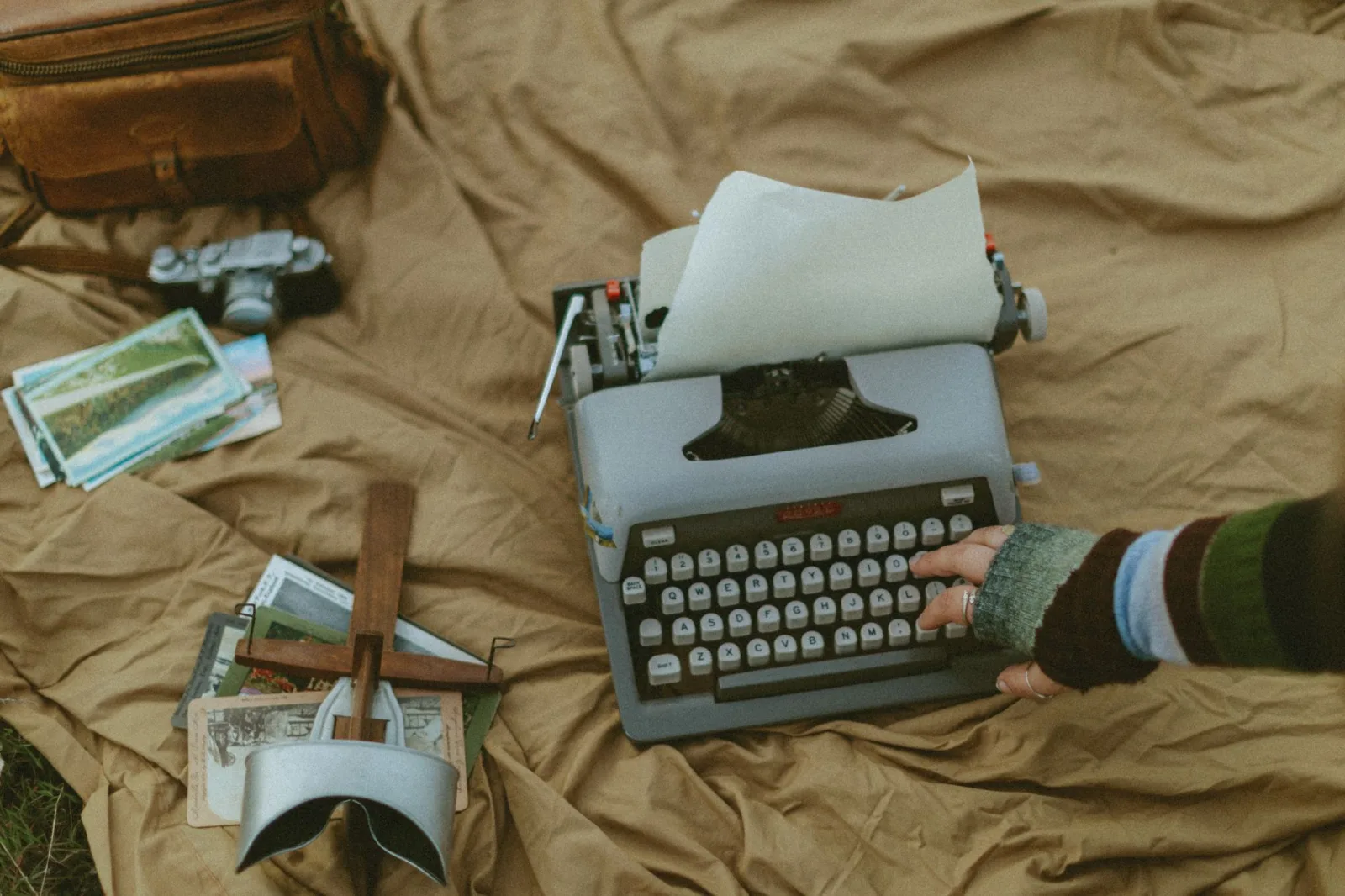 A person typing on a vintage typewriter outdoors, surrounded by postcards and a camera.