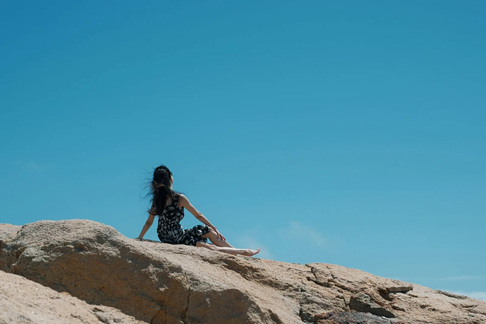 A woman enjoys solitude sitting on a rocky cliff under a bright blue sky, perfect for serene nature themes.
