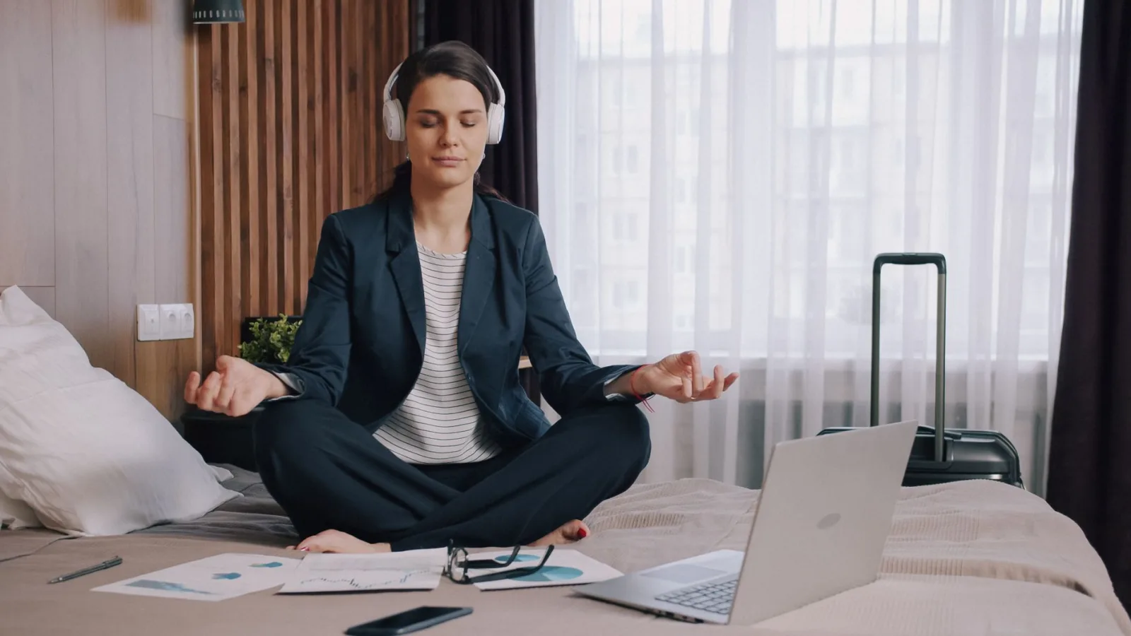 Businesswoman seated on a bed meditating with headphones and laptop in hotel room.