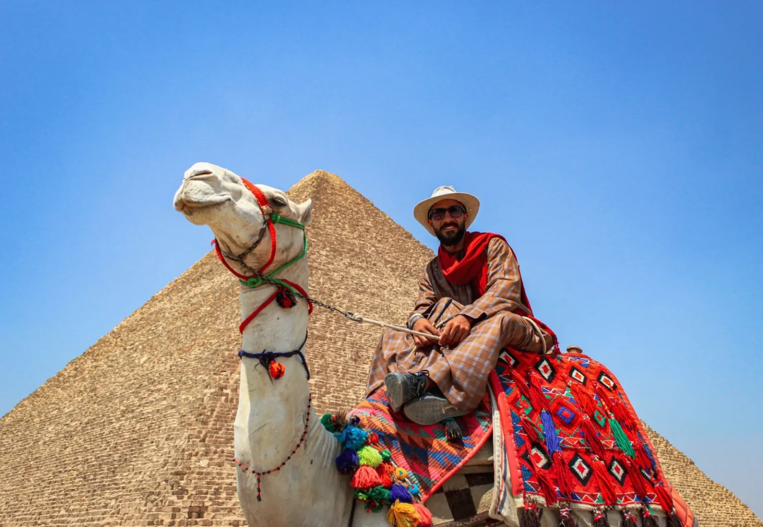 A man in traditional attire rides a camel near the Great Pyramid of Giza under a clear blue sky.