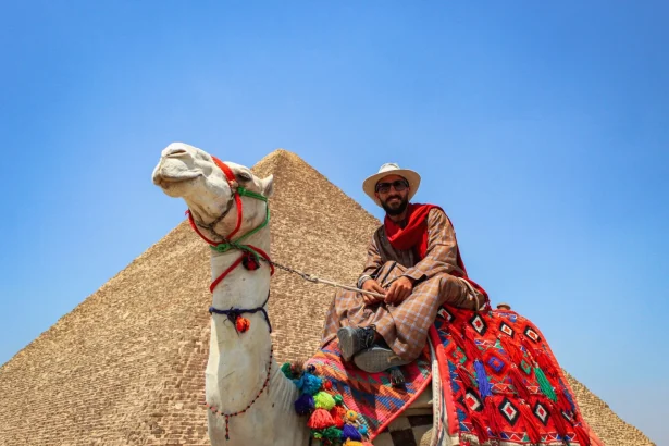 A man in traditional attire rides a camel near the Great Pyramid of Giza under a clear blue sky.