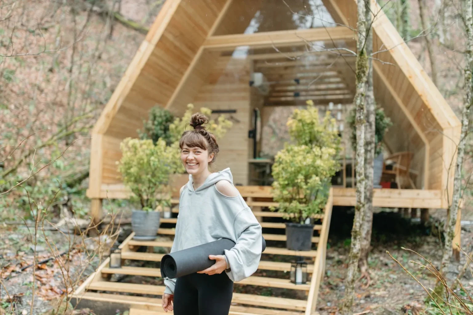 Cheerful woman with yoga mat in front of a wooden cabin surrounded by nature.