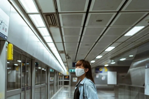 A woman wearing a face mask stands with luggage in a subway terminal, ready for travel.