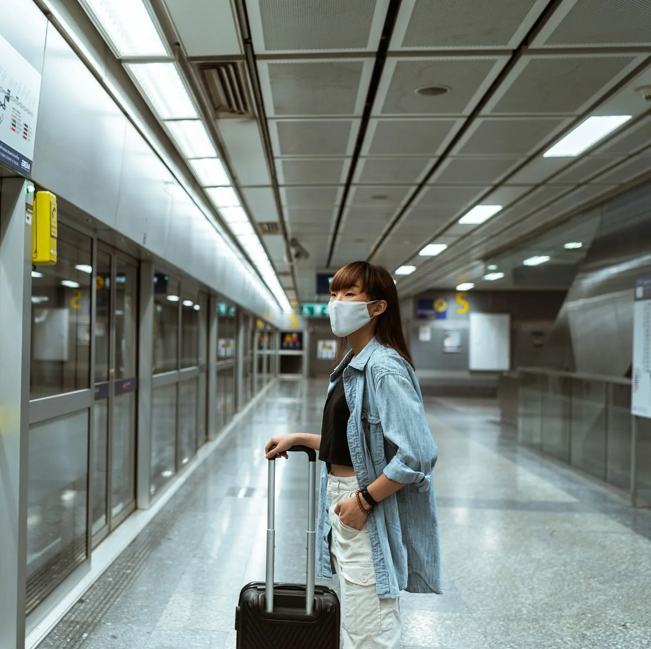 A woman wearing a face mask stands with luggage in a subway terminal, ready for travel.
