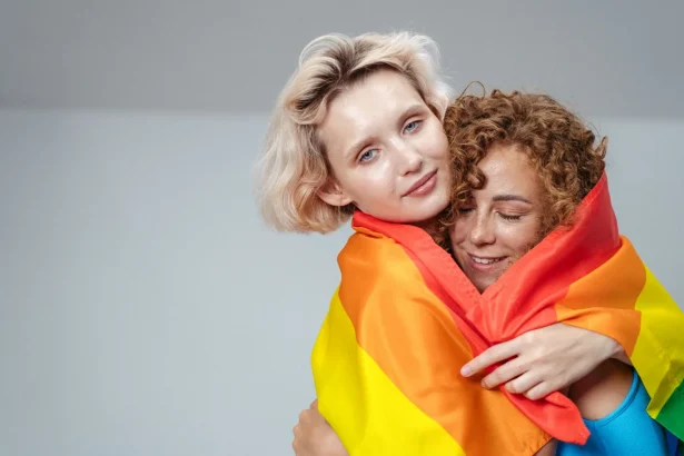 Two women lovingly embrace with a bright rainbow Pride flag, symbolizing LGBTQ love and support.