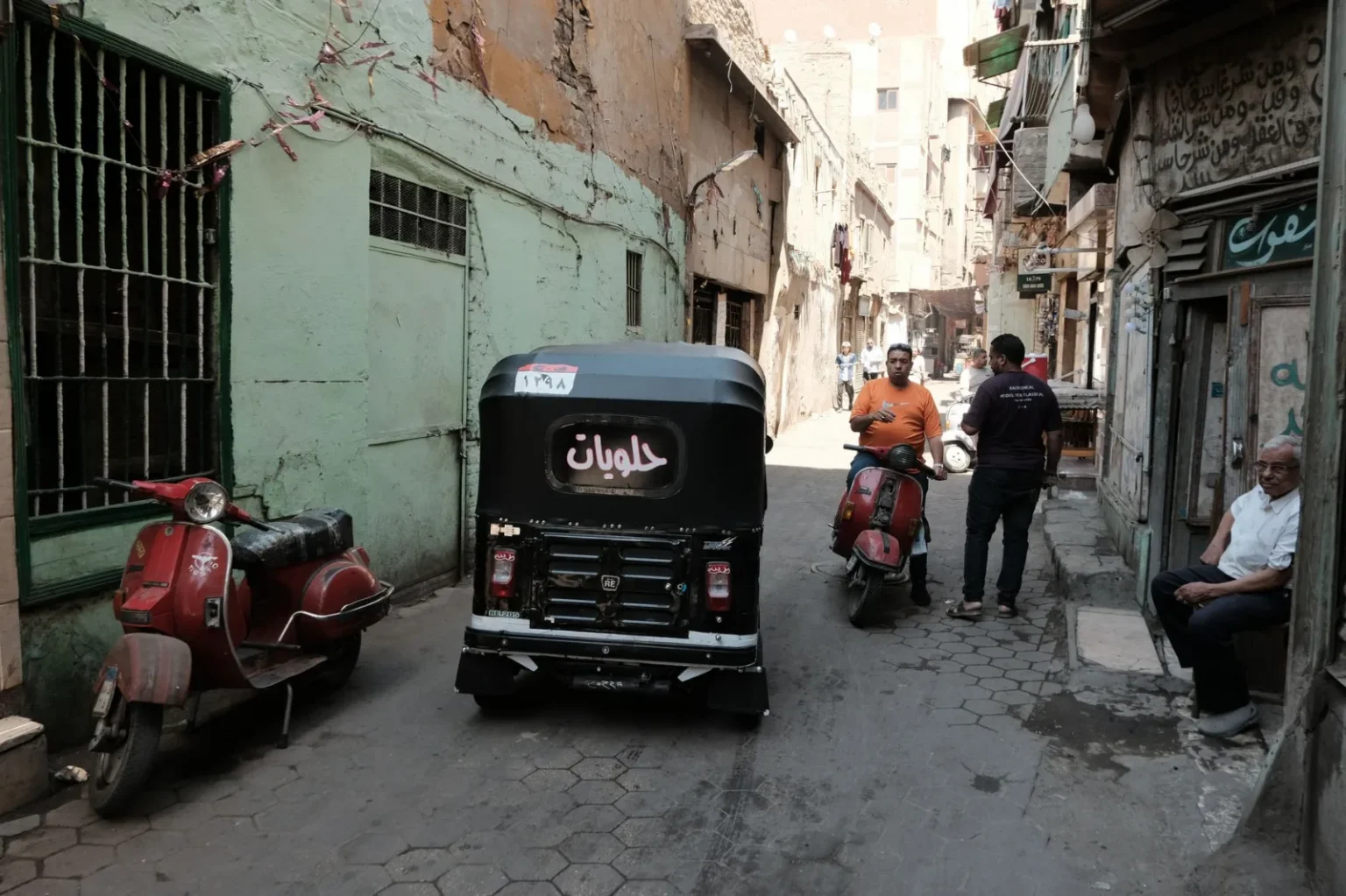 A bustling alleyway in Cairo featuring a tuk-tuk, scooters, and locals on a sunny day.