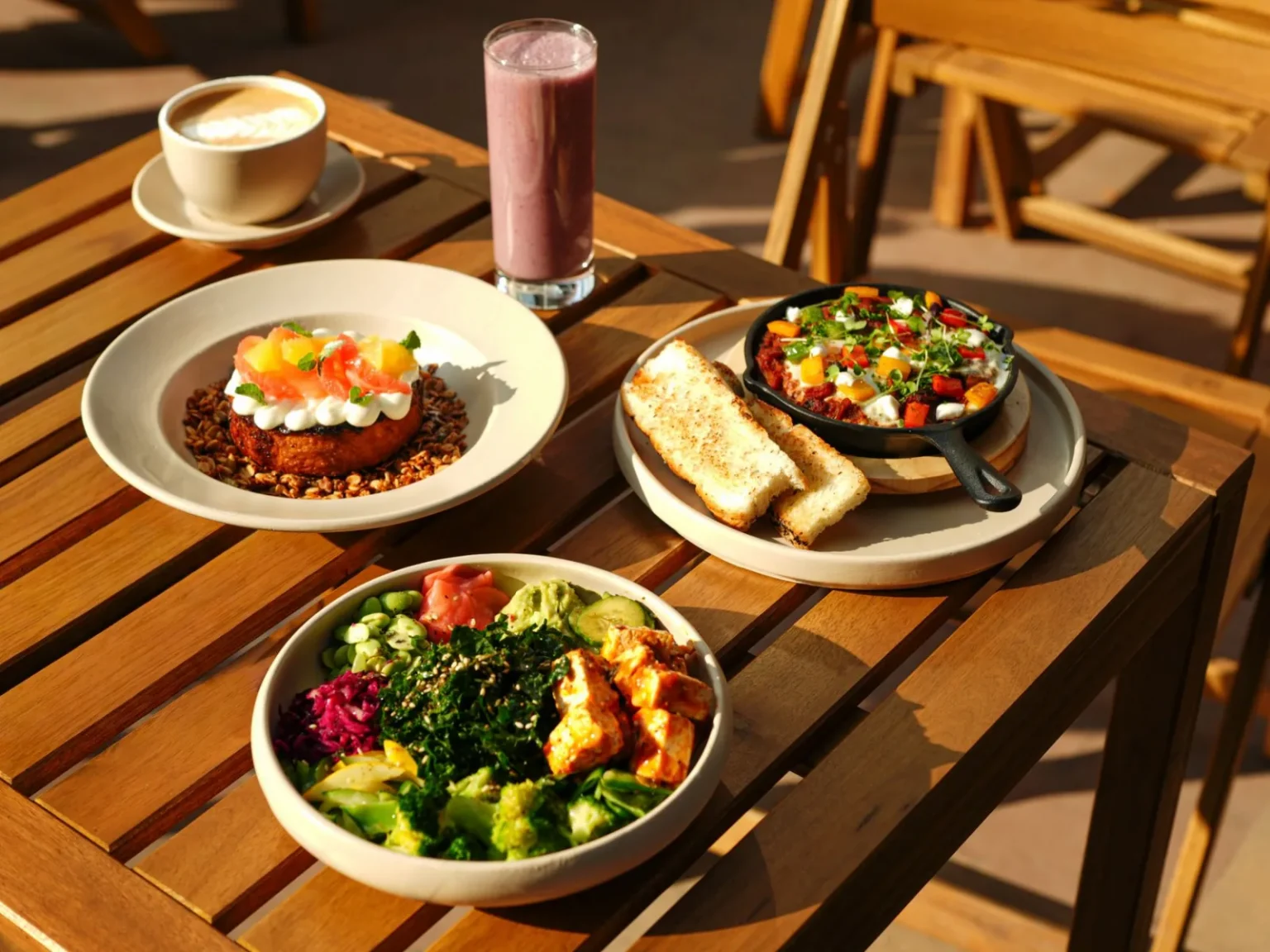 A vibrant set of healthy dishes including a smoothie and coffee on an outdoor wooden table.