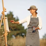 Cheerful woman in hat and apron painting while standing in summer sunny day on middle of field
