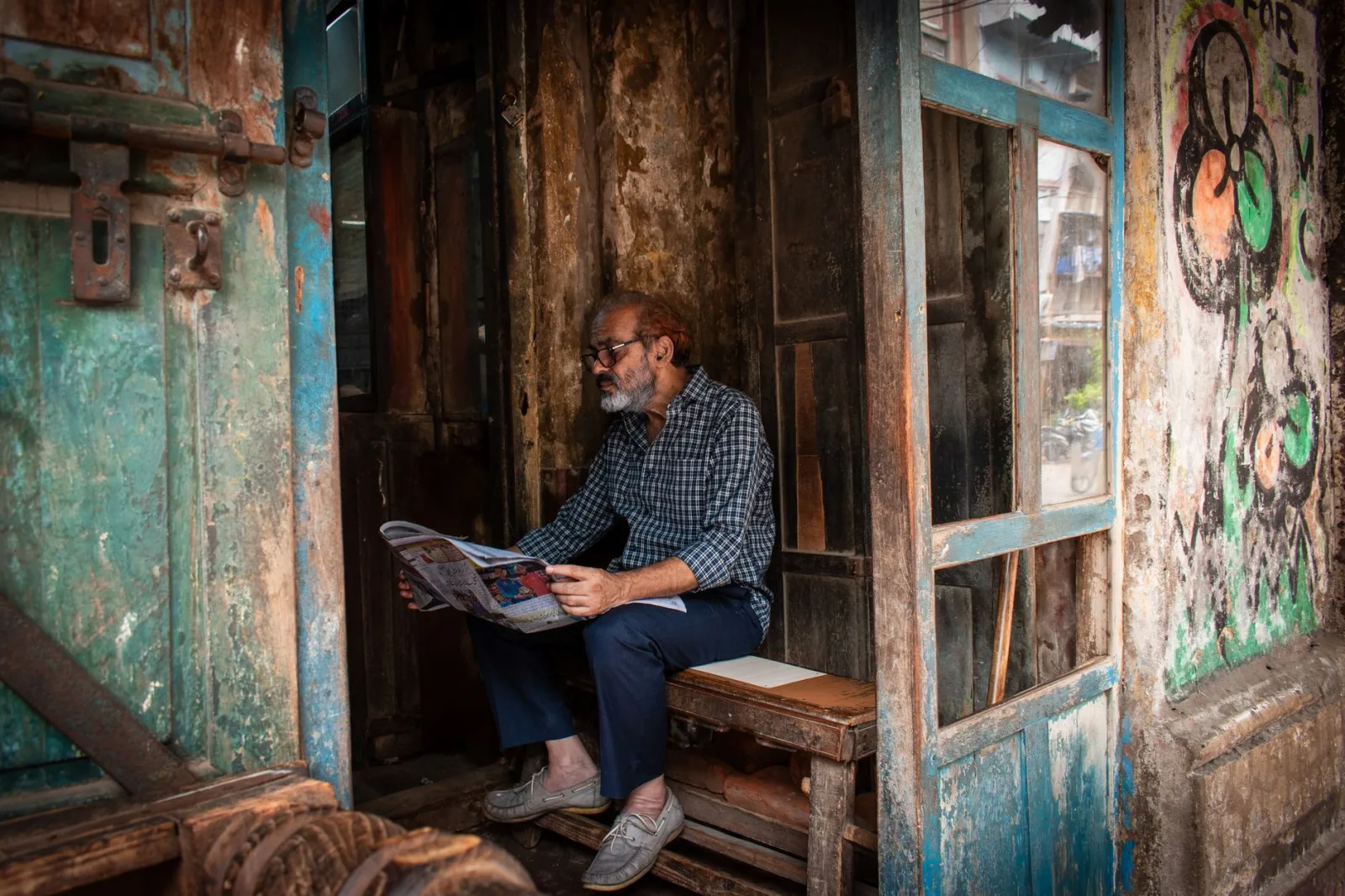Senior man sits in worn-out Kolkata alley reading a newspaper, surrounded by graffiti.