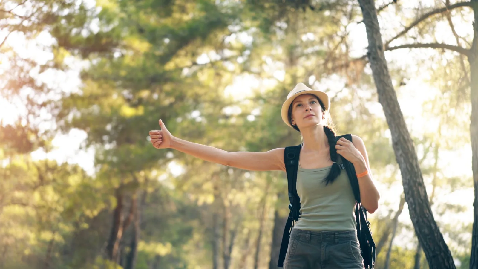 A woman with a backpack hitchhikes on a sunny day in the forest, wearing a hat.