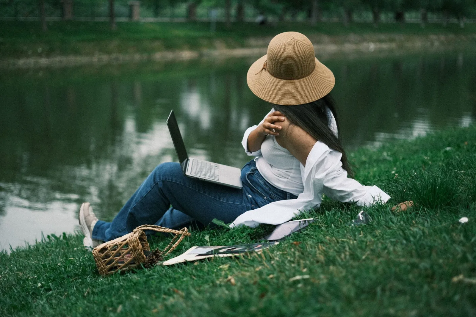 A woman relaxing by a lake using a laptop, enjoying outdoor work in a tranquil park environment.