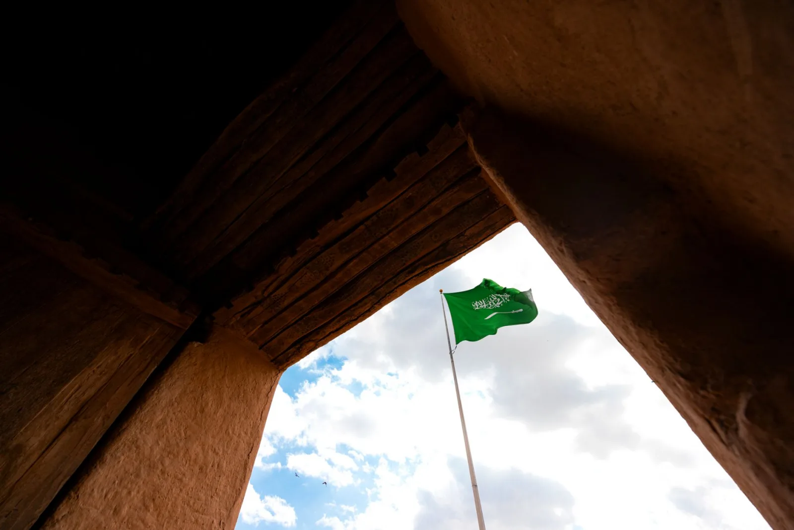 Saudi Arabian flag waves against blue sky amid traditional architecture.
