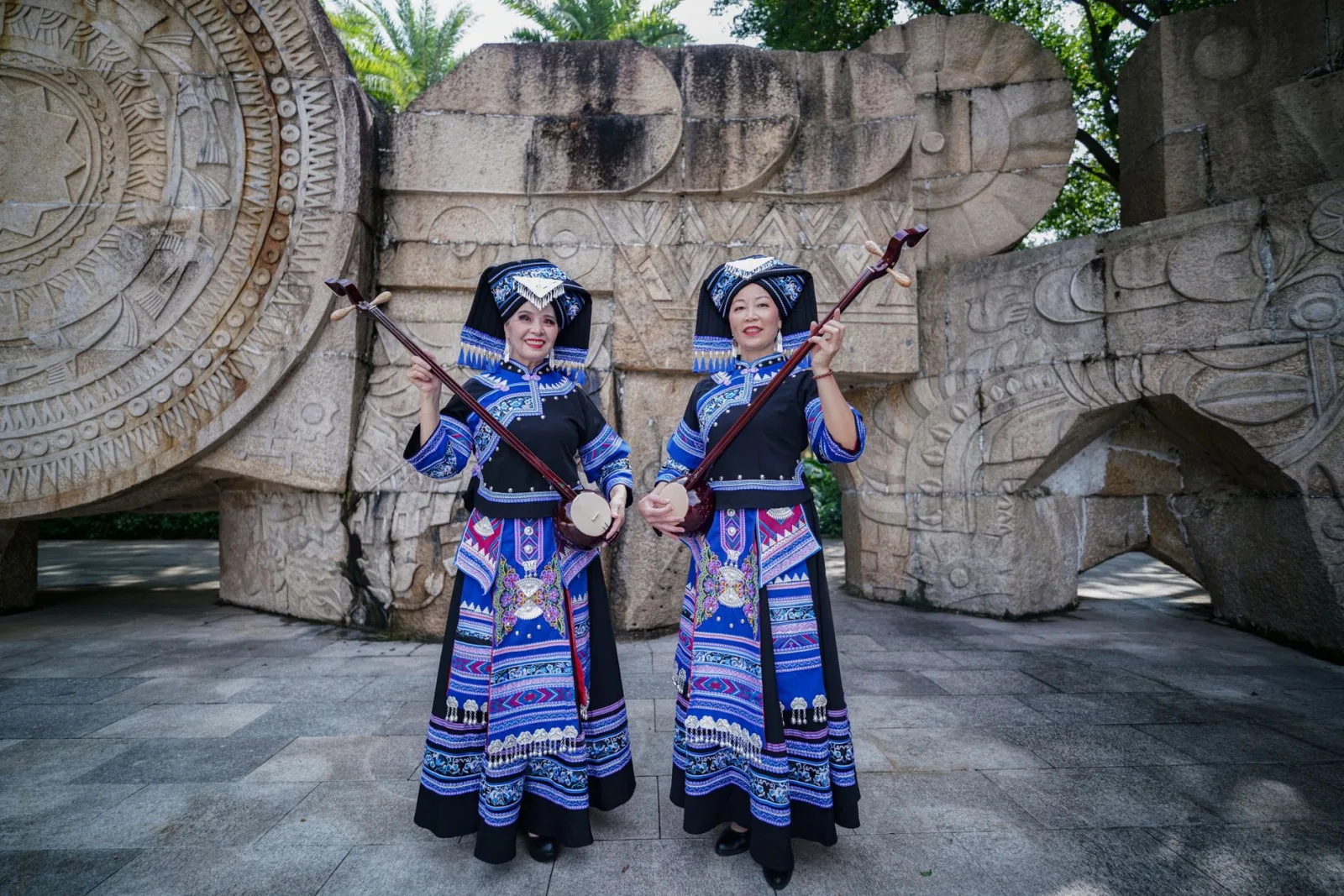 Two women in traditional Zhuang costumes playing musical instruments outdoors.
