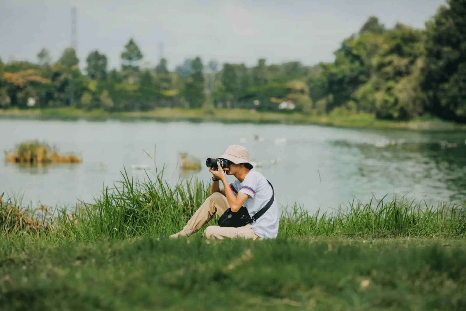 A young photographer sitting by a pond in a park capturing nature scenes on a summer day.