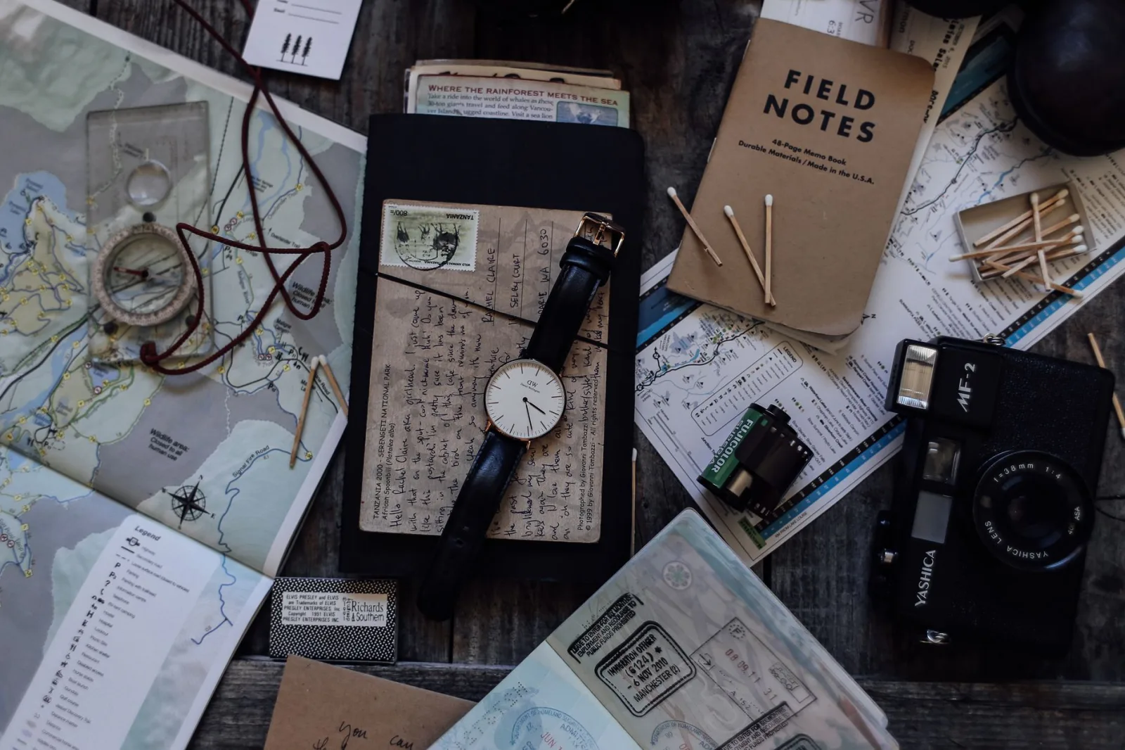 Top view of vintage photo camera near wristwatch and camera roll near maps and notebook near compass and papers near matches placed on table