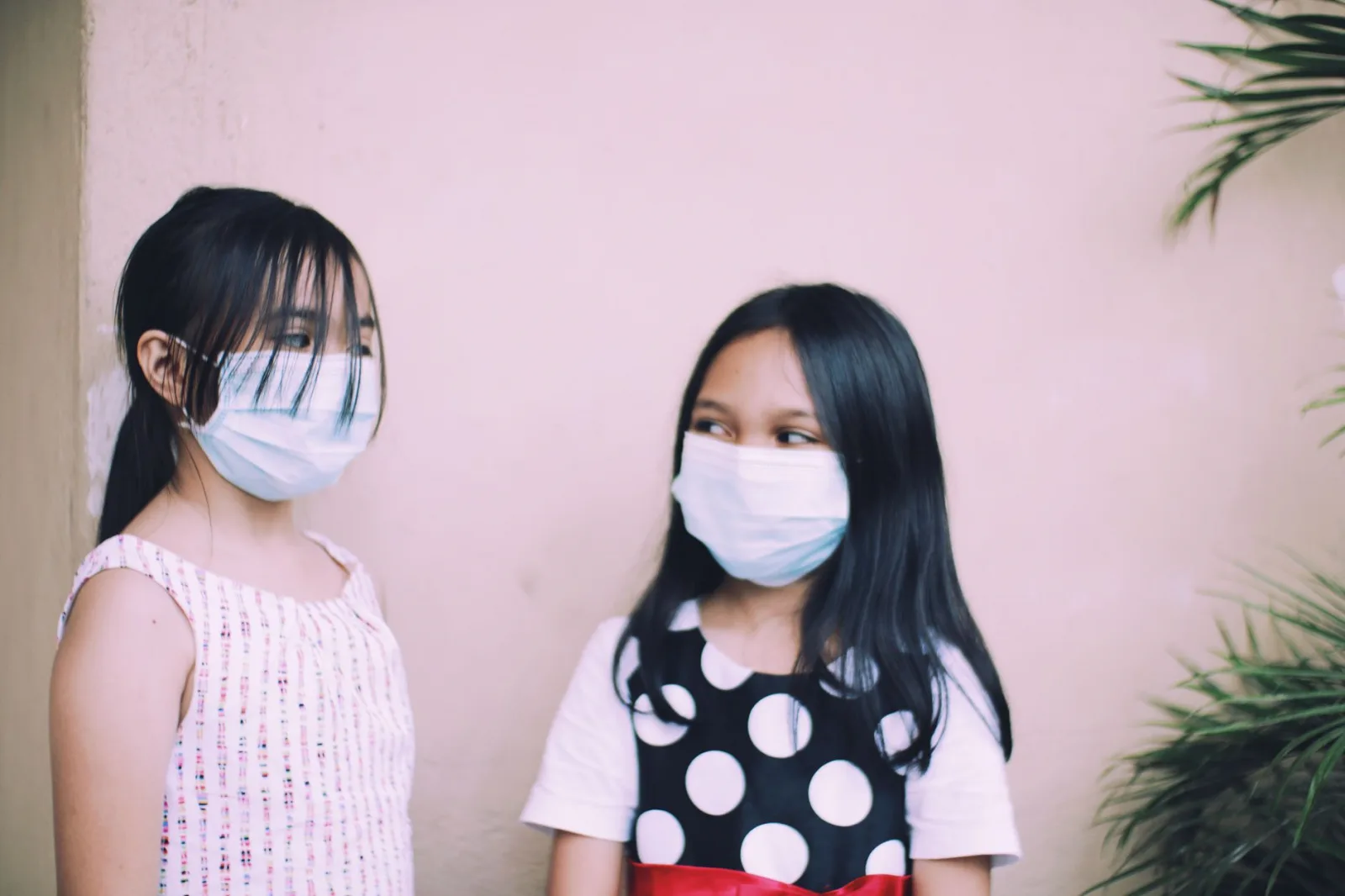 Two young girls in face masks engaging in conversation indoors, expressing friendship.