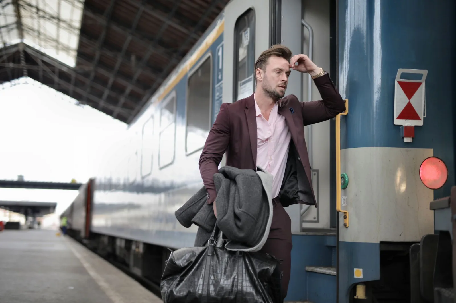 A man in a suit, carrying bags, looks stressed as he boards a train at a railway station.