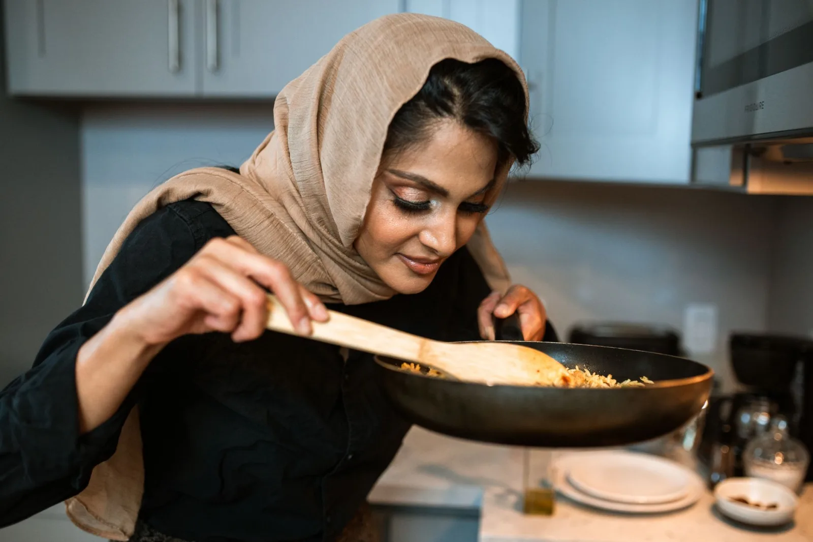 A Muslim woman wearing a hijab smiles while cooking with a frying pan in a modern kitchen.