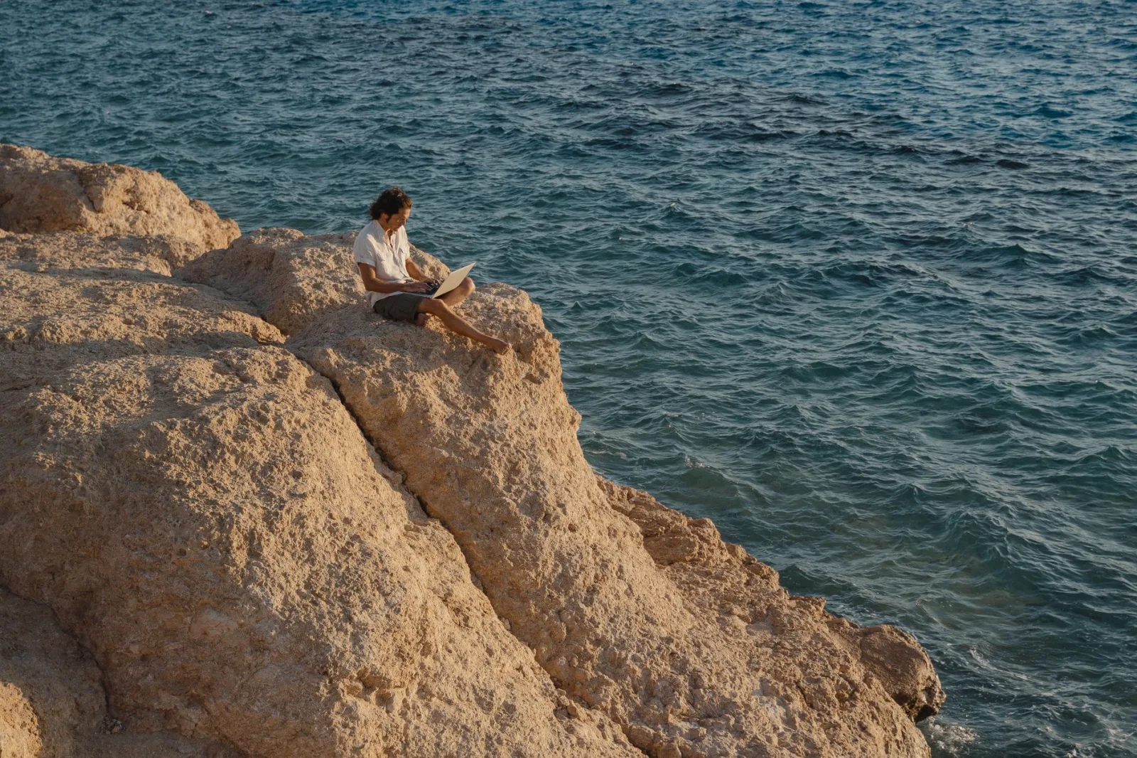 A man sits on a rocky cliff by the sea, working remotely on a laptop.
