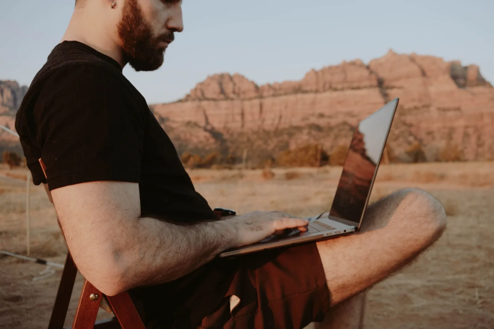 Side view of crop male freelancer sitting on chair while working on laptop in desert with mountain range