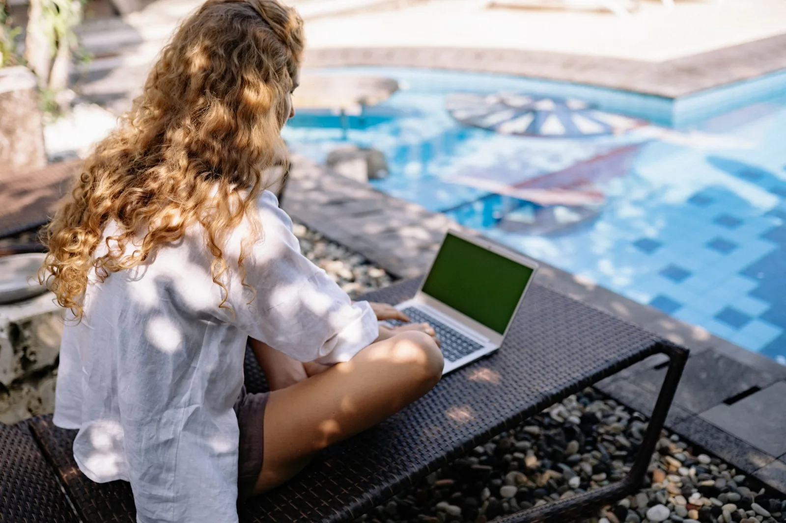 Woman with curly hair working on a laptop outdoors by a pool during summer, embodying a digital nomad lifestyle.