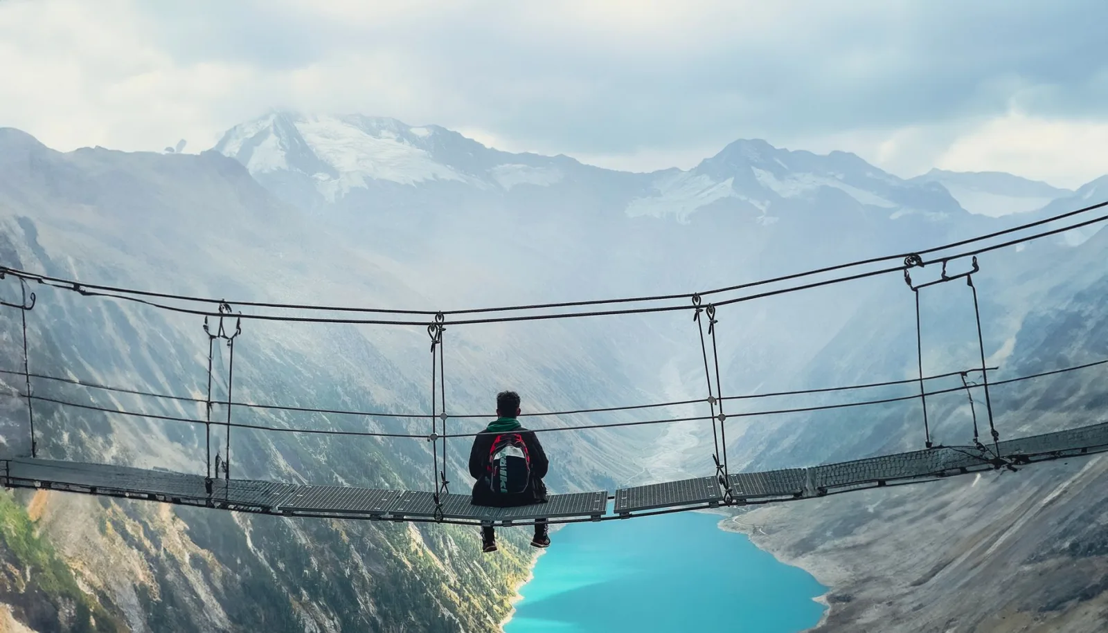A hiker sits on a suspension bridge overlooking a stunning alpine lake and snowcapped mountains.