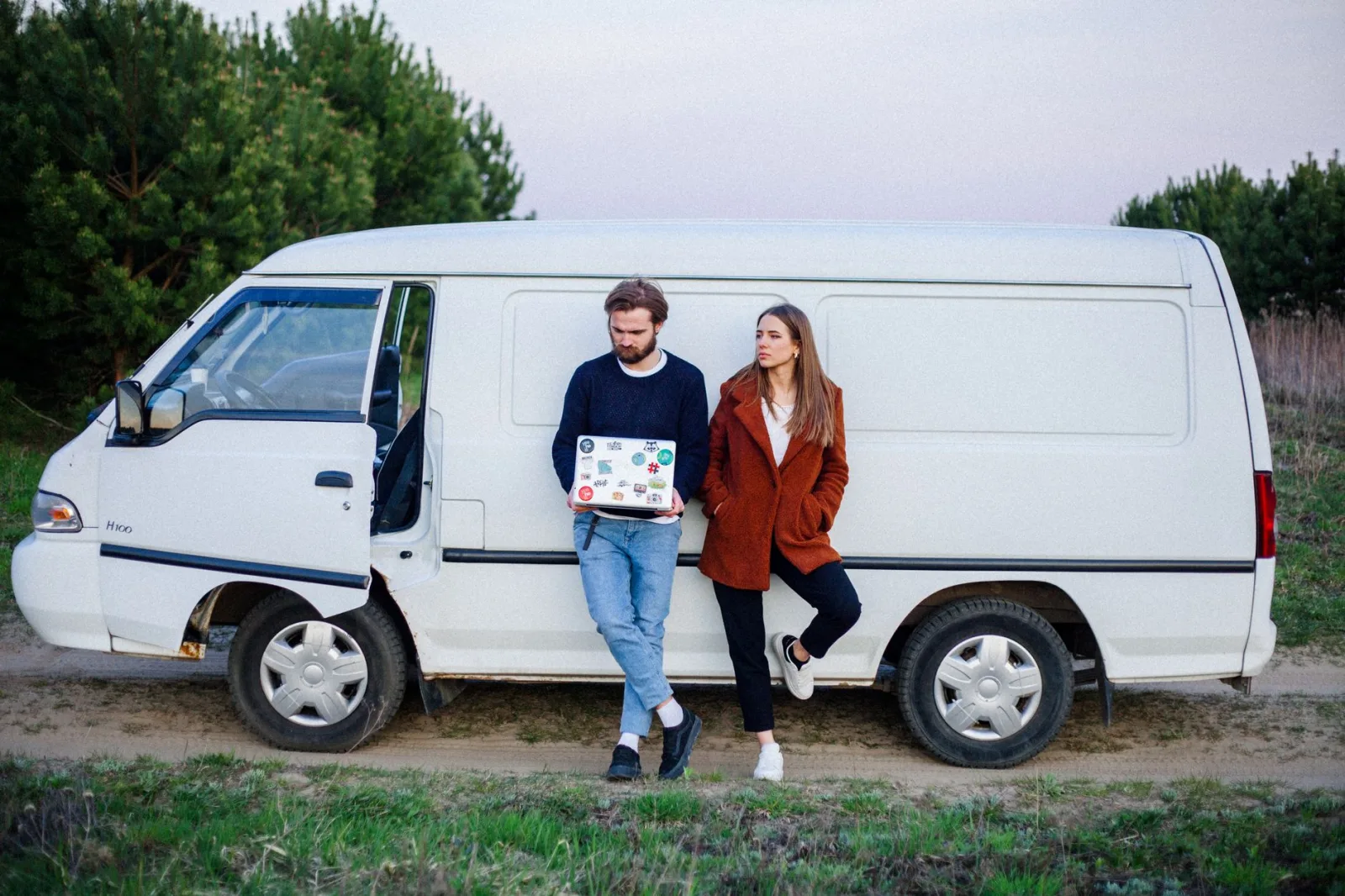 A couple leaning against a white van surrounded by greenery, enjoying a peaceful moment outdoors.