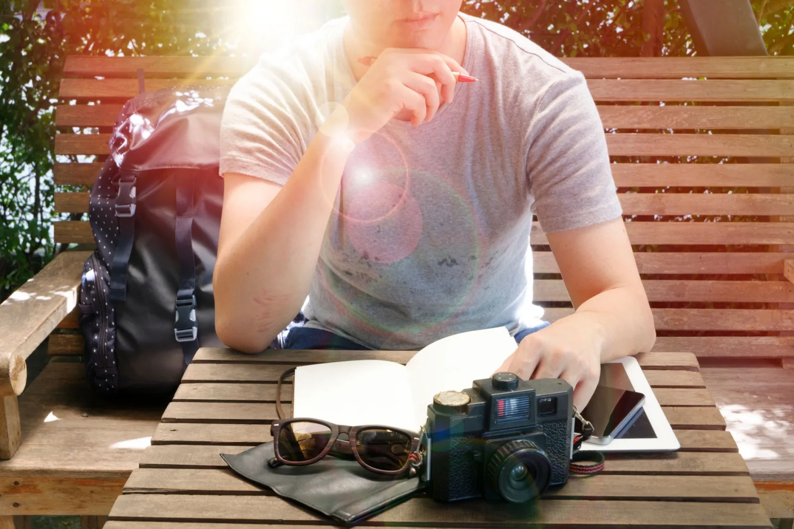 Man sitting on a wooden bench outdoors, reflecting with a camera, notebook, and sunlight surrounding him.