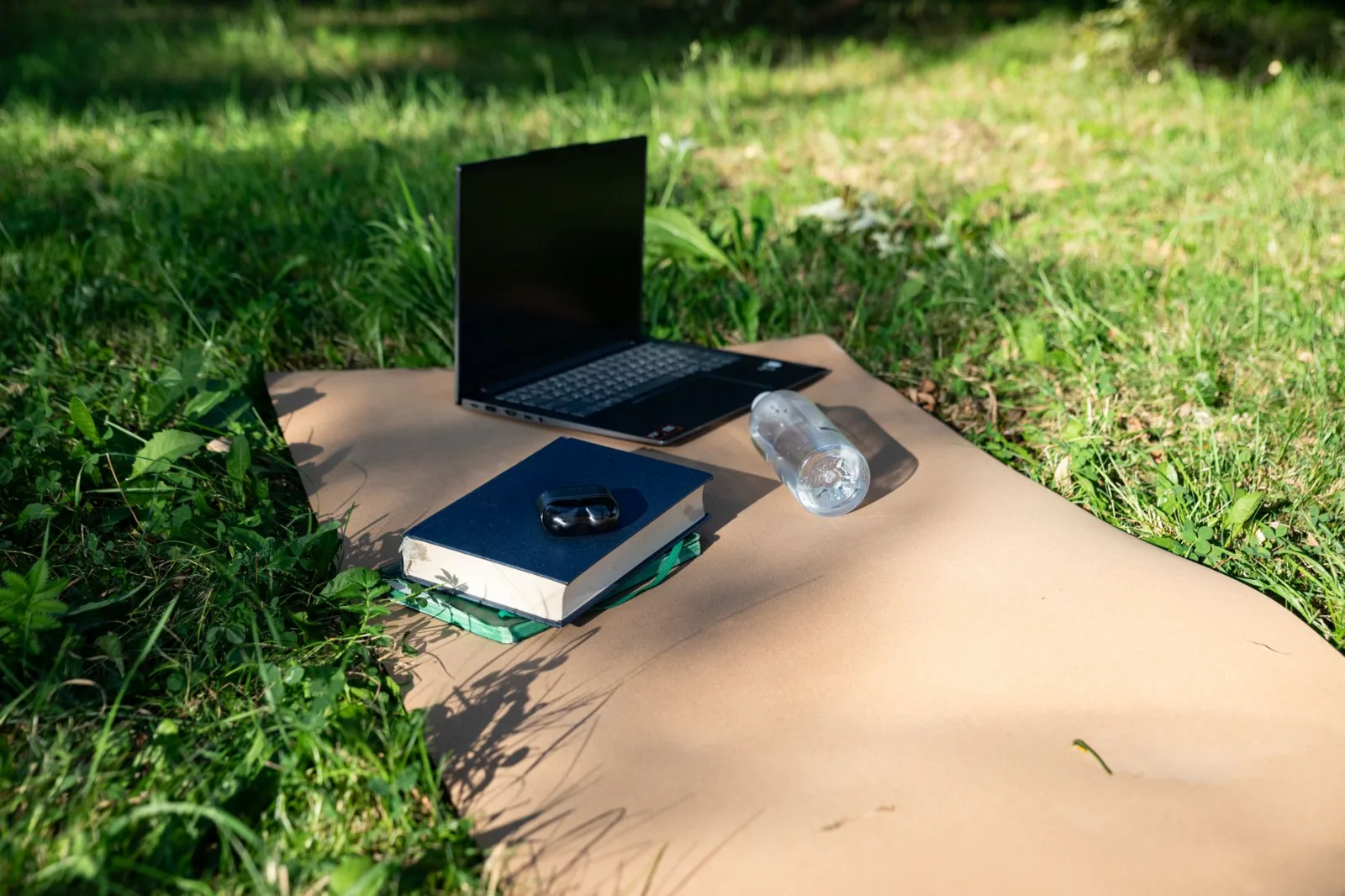 Laptop, books, and water bottle set on a mat in a sunny outdoor setting, ideal for remote work.