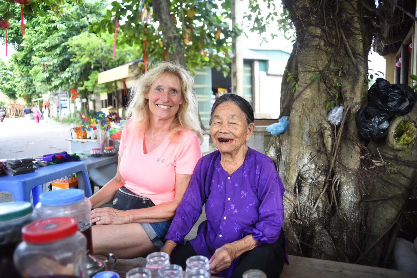 Two women of different backgrounds share a joyful moment at an outdoor market with trees and local goods.