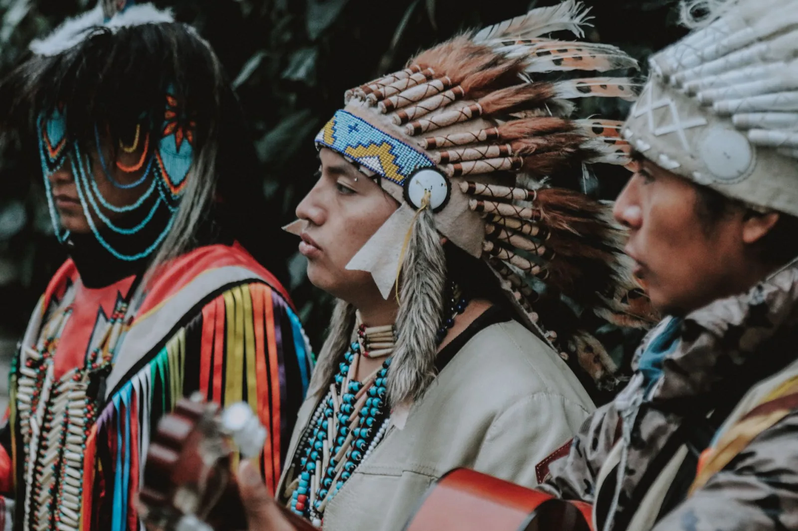 Three Native American men in traditional attire play guitars outdoors, capturing a cultural musical moment.
