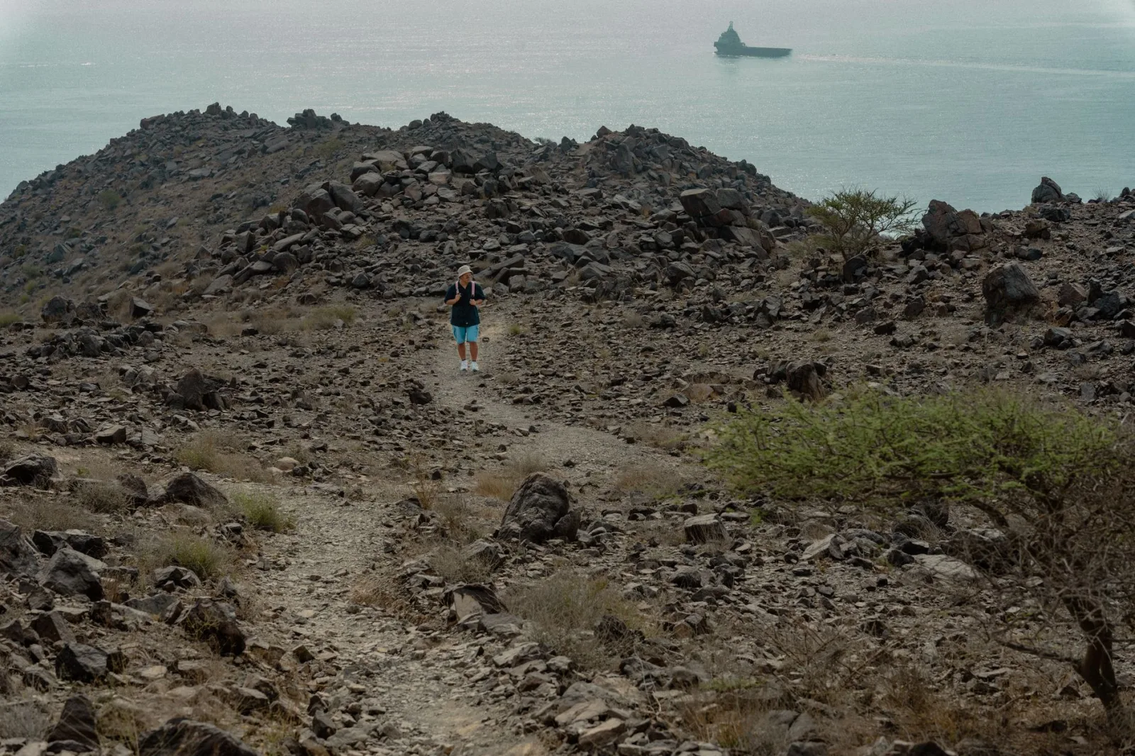 Hiker traversing rocky slopes with sea view in Khor Fakkan, UAE. Rugged mountain backdrop and tranquil waters.