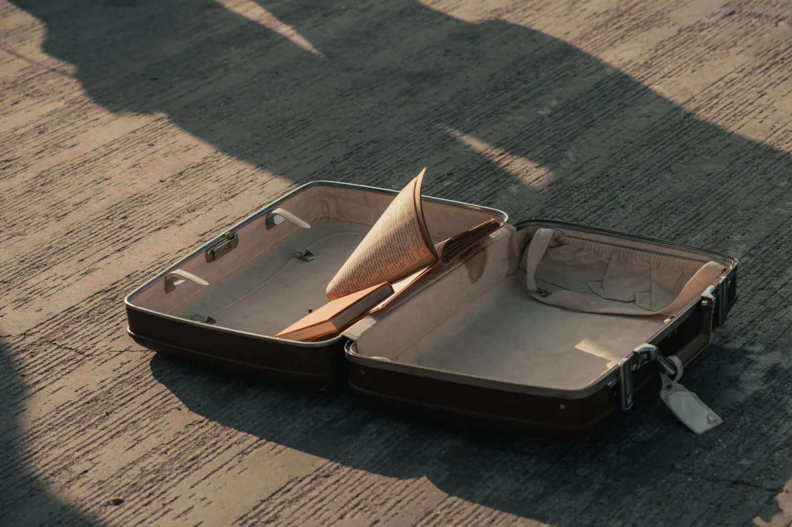 An open suitcase on a pavement with a book inside, casting shadows.