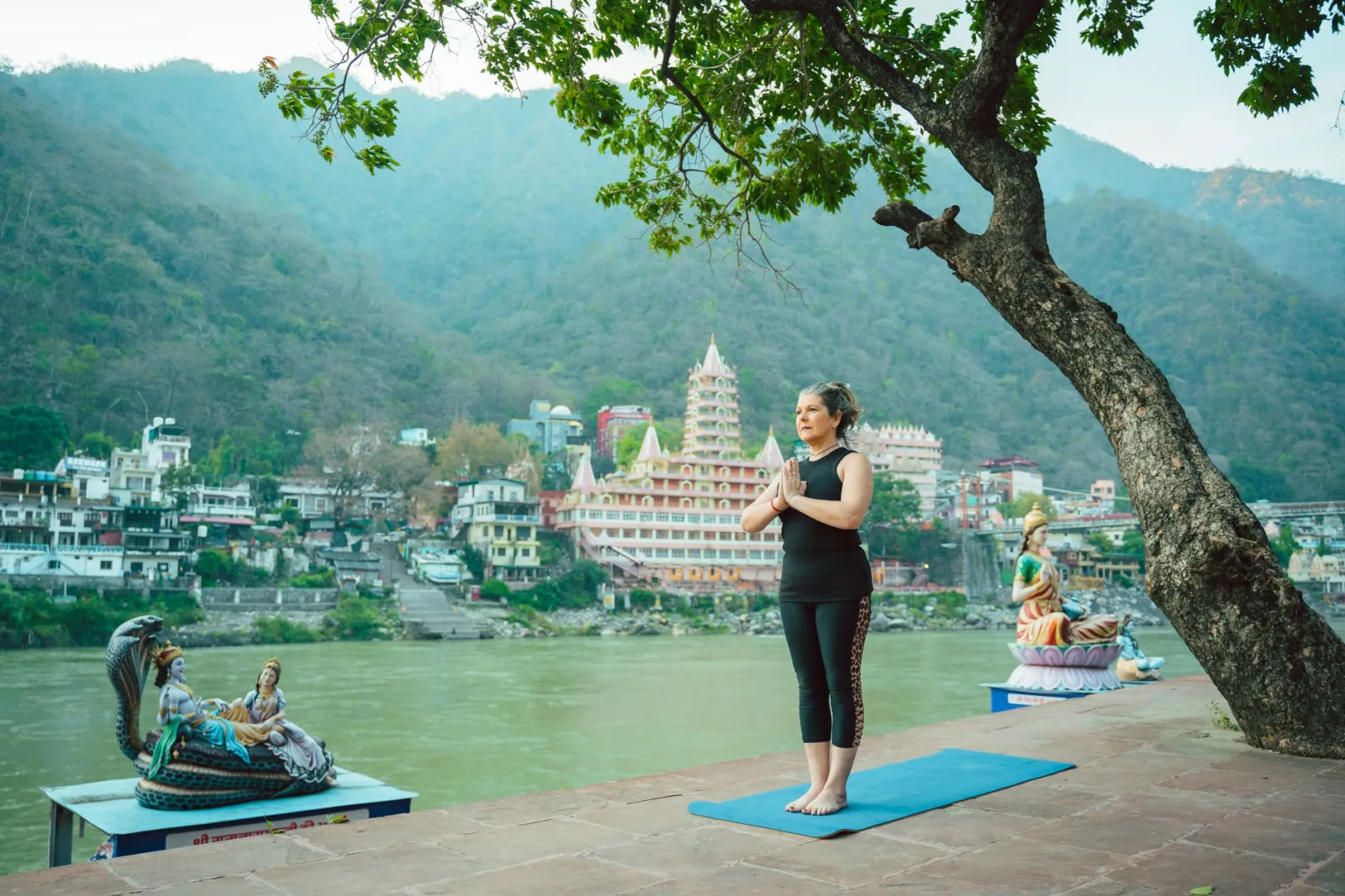 Woman practicing yoga by a serene riverside with statues in the backdrop, Rishikesh, India.