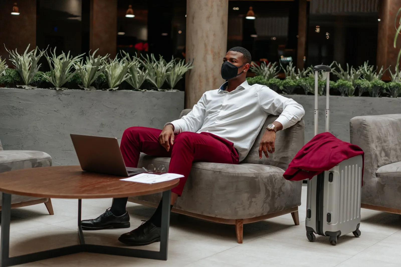 Man wearing facemask, sitting in hotel lobby with laptop and suitcase, adapting to new normal.