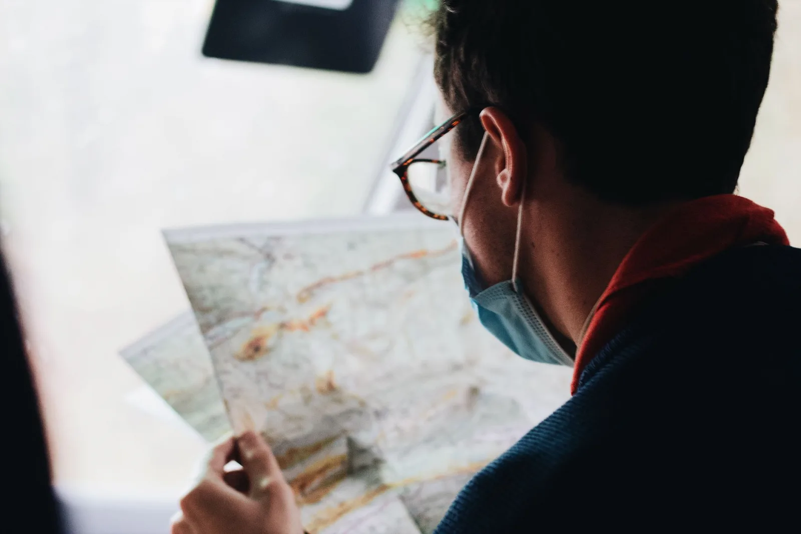 A man wearing a face mask examines a map while traveling in a vehicle during daytime.