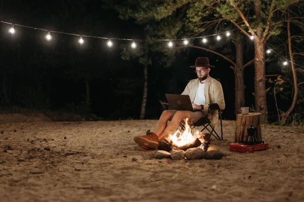 Digital nomad working remotely by a campfire with a laptop under string lights in the forest.