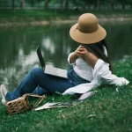 A woman relaxing by a lake using a laptop, enjoying outdoor work in a tranquil park environment.