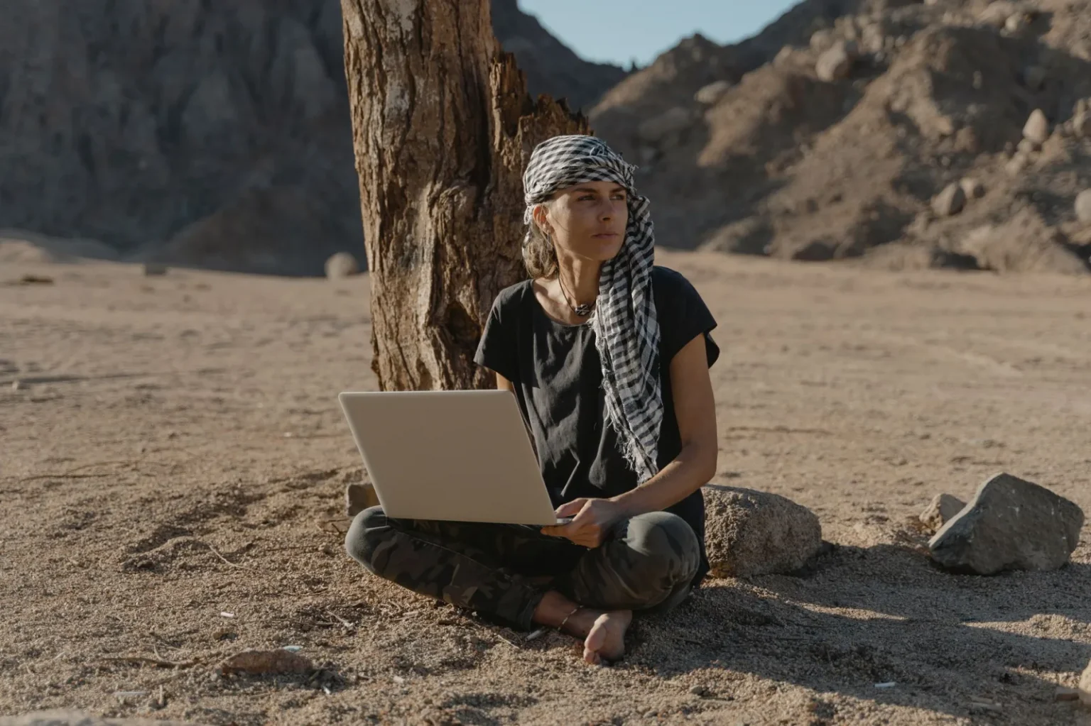 Caucasian woman sitting barefoot with laptop under tree in arid landscape.