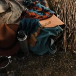 Close-up view of camping essentials including a thermos, backpack, and hat on a blanket.