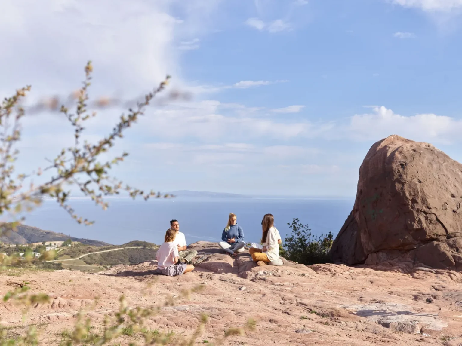 Four adults meditate on a rocky cliff overlooking the ocean under a clear, sunny sky.
