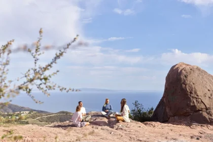Four adults meditate on a rocky cliff overlooking the ocean under a clear, sunny sky.
